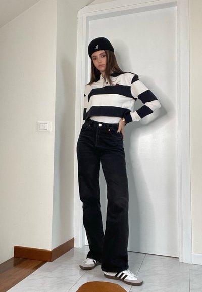 Polo a rayas blanco y negro, jeans negros de tiro alto y zapatillas negras con rayas blancas. Llevado con un gorro negro, de pie en el interior.