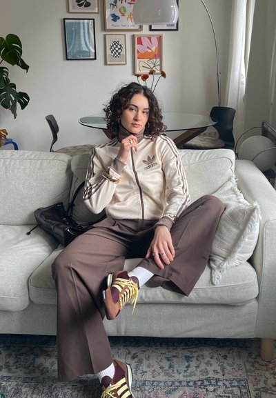 Mujer joven con cabello rizado que lleva una chaqueta beige de Adidas y pantalones anchos marrones, sentada en un sofá gris claro en una sala de estar con arte en las paredes y plantas.