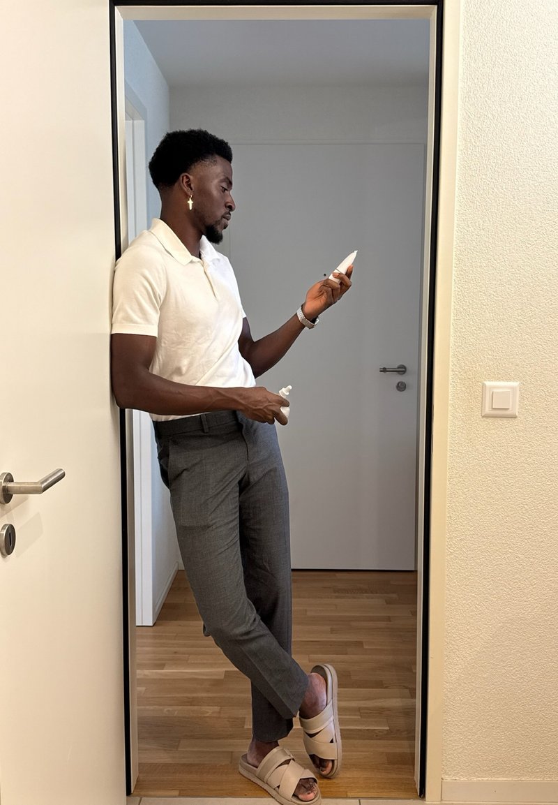 Un homme portant un polo blanc et un pantalon gris se tient dans une porte, tenant des produits de soin de la peau. Il porte des sandales beiges et des accessoires en argent.