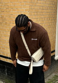 Hnědý fleecový pulovr s límcem a bílou výšivkou, doplněný černými kalhotami a krémovou crossbody kabelkou. Splétané vlasy a brýle.