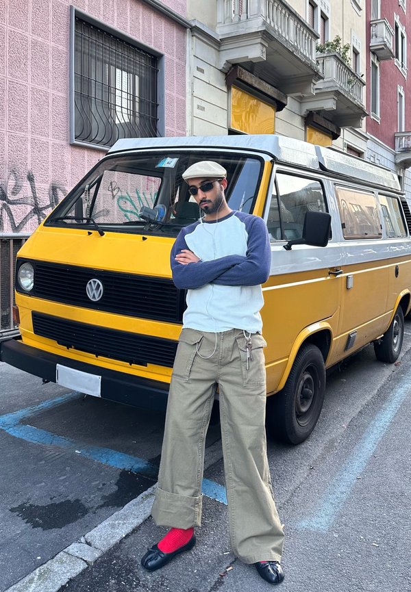 Homme bras croisés portant un chapeau, des lunettes de soleil et des écouteurs, se tient à côté d'un van Volkswagen jaune et blanc dans une rue de la ville.