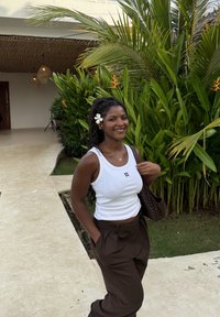Femme souriante aux longs cheveux avec une fleur blanche, vêtue d'un débardeur blanc et d'un pantalon marron, debout à côté de plantes tropicales vertes en plein air.