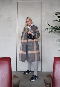 Manteau à carreaux gris et orange porté sur un hoodie gris, associé à des leggings gris et des chaussures de sport noires. Petit sac à main noir avec des accents de chaîne.
