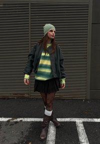 Šedá bomber bunda přes svetr s barevnými pruhy, černá sukně, silonkové punčocháče s puntíky a hnědé kozačky po kolena. Zelená pruhovaná čepice.