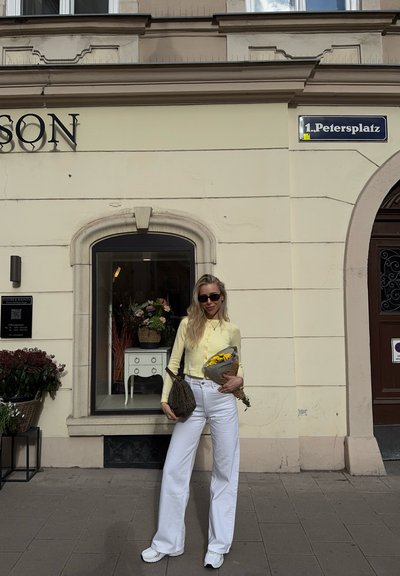 Una mujer se encuentra frente a una tienda, vistiendo una camisa amarilla de botones, pantalones blancos de pierna ancha, sosteniendo un ramo de flores y un bolso marrón con estampado.