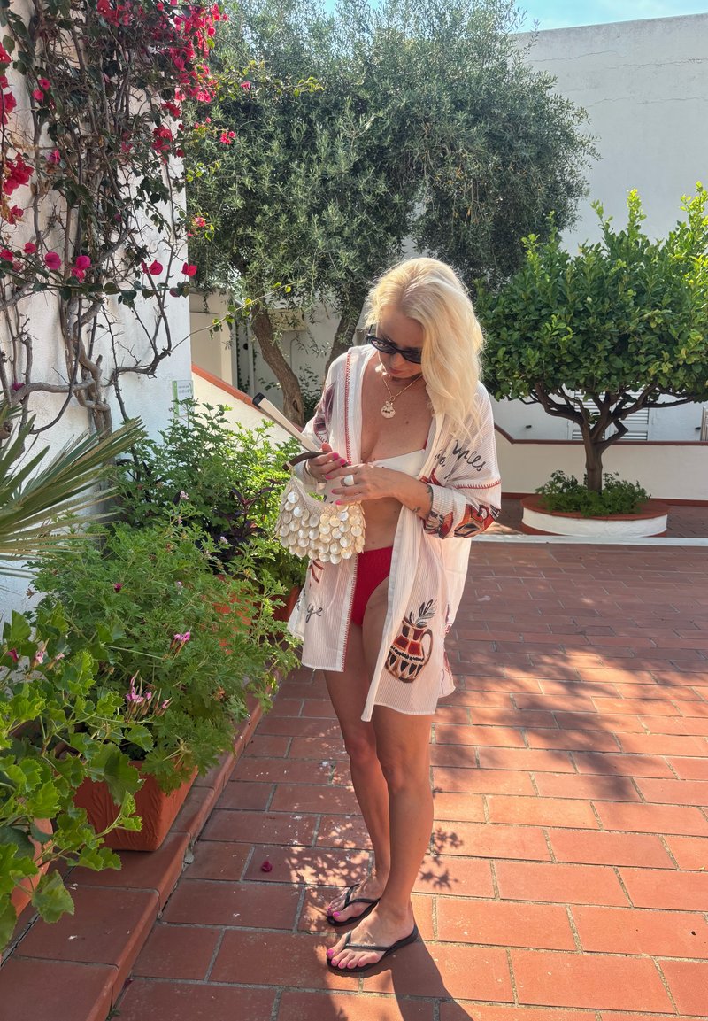 Femme blonde en lunettes de soleil et tenue de plage, debout sur une terrasse carrelée entourée de plantes en pot et d'arbres, tenant un petit sac.