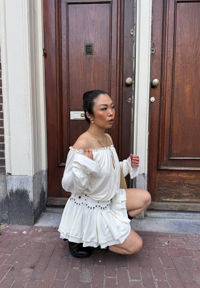 Vestido blanco de tirantes caídos con falda de volantes y detalles de ojales decorativos. Lleva botas negras y está de pie junto a puertas de madera con herrajes de metal.