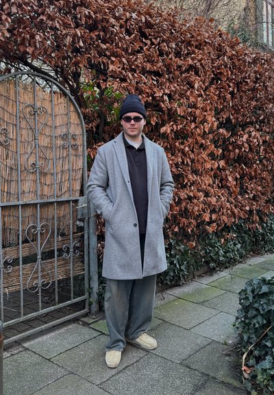 Abrigo de lana gris con un corte relajado, llevado sobre una camiseta polo oscura y pantalones holgados. Un gorro negro y gafas de sol completan el atuendo.