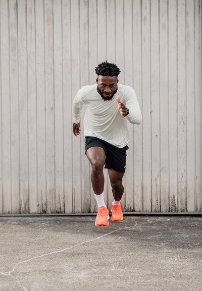 Atleta con una camiseta de manga larga blanca y pantalones cortos negros, corriendo. Zapatillas de deporte naranjas brillantes, calcetines blancos, fondo de concreto texturizado.
