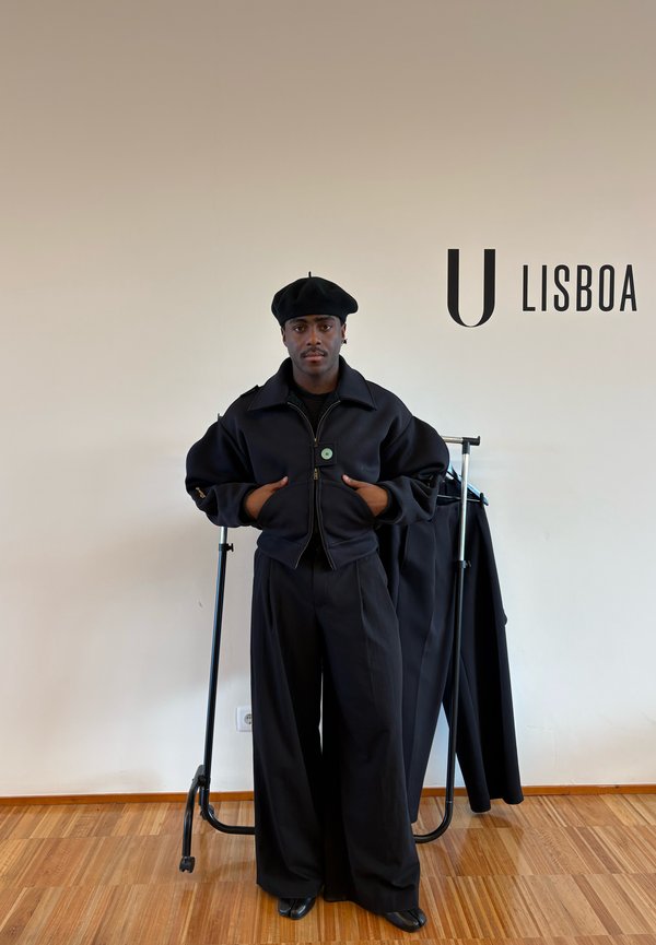 Mann mit schwarzer Baskenmütze und übergroßem schwarzem Outfit steht drinnen neben einem Kleiderständer mit schwarzer Kleidung, mit dem Text "U Lisboa" an der beigen Wand im Hintergrund.