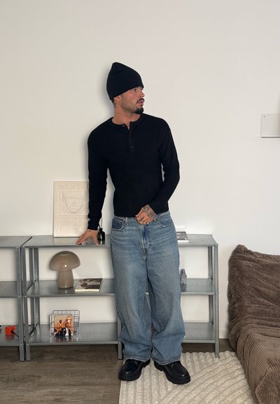 Camisa henley negra de manga larga, vaqueros holgados azul claro, gorro negro. De pie cerca de una estantería metálica con objetos decorativos y una manta marrón con textura.