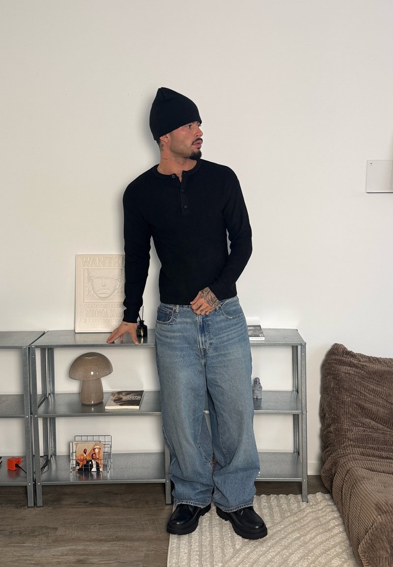 Chemise henley noire à manches longues, jean ample bleu clair, bonnet noir. Debout près d'une étagère métallique avec des objets décoratifs et une couverture marron à texture.