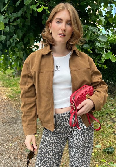 Chaqueta de ante marrón, top corto blanco con logo, pantalones de leopardo, bolso acolchado rojo con herrajes dorados, llevando gafas de sol.