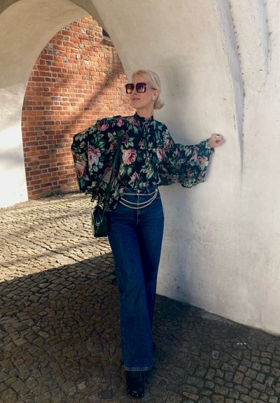 Blusa floral de manga larga con mangas acampanadas en verde oscuro, combinada con jeans anchos de talle alto en azul y botas de plataforma negras. Bolsa negra con cadena.