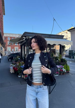 Mujer con cabello rizado ajusta una chaqueta negra sobre una camisa a rayas mientras está cerca de un puesto de flores en una calle de la ciudad bajo un cielo despejado.