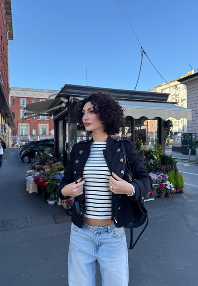 Mujer con cabello rizado ajusta una chaqueta negra sobre una camisa a rayas mientras está cerca de un puesto de flores en una calle de la ciudad bajo un cielo despejado.