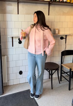 Suéter rosa y peludo con capucha, jeans de campana azul claro y zapatillas negras. Sosteniendo una bebida, apoyada contra una pared de azulejos.
