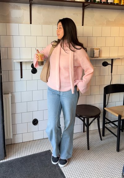 Suéter rosa y peludo con capucha, jeans de campana azul claro y zapatillas negras. Sosteniendo una bebida, apoyada contra una pared de azulejos.
