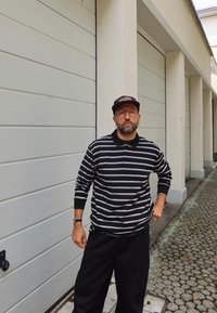 Mężczyzna z brodą, w okularach i czapce, ubrany w czarno-białą pasiastą koszulę z długim rękawem i czarne spodnie, stojący na brukowanej drodze przy garażach.