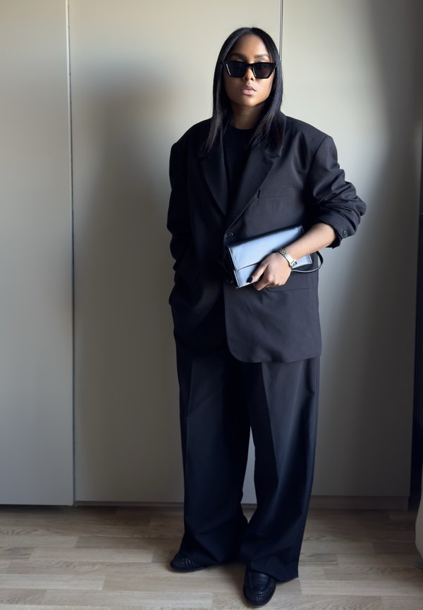 Sort oversized blazer, sort top og vidde bukser. Iført sorte solbriller og holder en blå clutch. Glat stof, minimalistisk design.