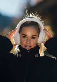 Młoda kobieta poprawiająca białe, puszyste nauszniki, ubrana w czarny płaszcz z wysokim kołnierzem, na zewnątrz, w miękkim świetle słonecznym, na rozmytym tle.