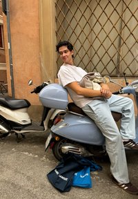 Blå scooter med et brunt sæde, en hvid t-shirt, lyse jeans, og to tote bags på jorden. Baggrunden har en beige væg og et gittervindue.