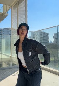 Chaqueta negra de rayas con cremallera al frente, con un parche de logo, combinada con una camiseta sin mangas blanca y pantalones a juego de rayas. Ambiente en un balcón de hormigón.