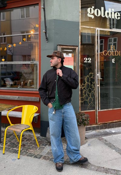 Chaqueta ligera negra, camisa verde, vaqueros holgados azul claro, zapatos marrones, gorra de cuadros. Silla metálica amarilla y planta cerca, fondo de tienda.