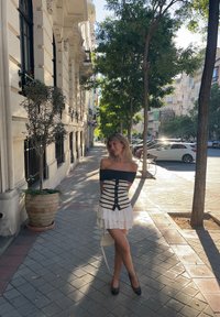 Topless φόρεμα με μαύρες και λευκές ρίγες, ρυακιωτή κοντή φούστα, κουμπιά και εφαρμοστό κορσέ; συνδυασμένο με μαύρες γόβες και μια μικρή λευκή τσάντα.