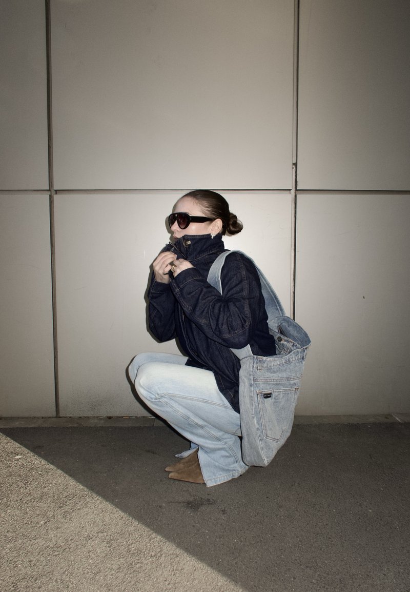 Person som sitter på huk utendørs, iført solbriller, en mørk jakke glidelåst opp, lyse jeans og bærer en stor denimveske mot en flislagt vegg.