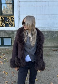 Smeđa jakna s obrubom od krzna i teksturiranim vunenim tijelom, s tamnim gumbima i krojenim oblikom, uparena sa sivim džemperom i crnim hlačama.