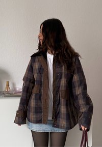 Jeune femme aux longs cheveux foncés portant une veste à carreaux par-dessus une chemise blanche et une jupe en denim, tenant un sac à main sombre, debout à l'intérieur.