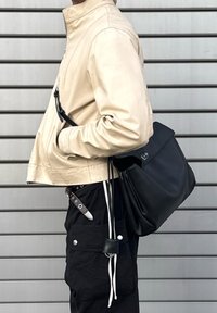 Béžová kožená bunda, zkrácený střih se stojáčkovým límcem. Černá taška přehozená přes rameno, vedle černých kalhot s kapsami a kovovými detaily.