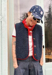 Personne souriante, portant une casquette à motifs d'étoiles, des lunettes de soleil, un bandana rouge, un gilet bleu marine avec bordure rouge, un t-shirt blanc et un pantalon sombre plissé.