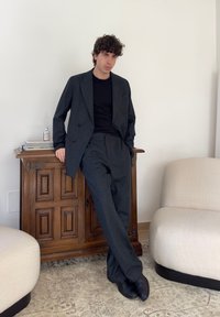 Junger Mann in dunkelgrauem Anzug und schwarzem Hemd lehnt an einem Holzschrank zwischen zwei beigen Sessel in einem minimalistischen Raum.