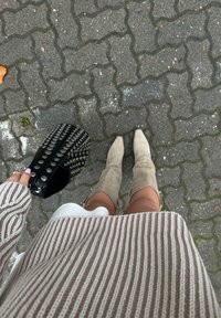 Tan suède kniehoge laarzen, een beige en crèmekleurige gestreepte trui, en een zwarte studded handtas op een gestructureerde grijze stoep.