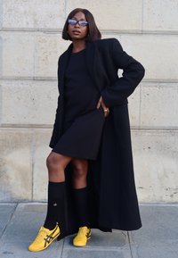 Schwarzer Mantel über einem schwarzen Kleid, kombiniert mit knielangen schwarzen Socken und leuchtend gelben Sneakers. Ausgestattet mit eleganten Sonnenbrillen und einem strukturierten steinigen Hintergrund.