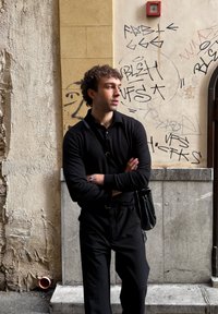 Homme en tenue noire avec un sac en bandoulière, les bras croisés, se tenant devant un mur couvert de graffiti dans un environnement urbain.