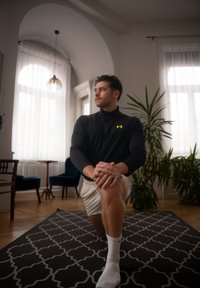 Homme portant un haut de sport noir zippé avec un logo jaune, à genoux sur un tapis à motifs, vêtu de shorts blancs et de chaussettes, dans un cadre intérieur avec des plantes.