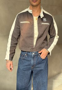 Brun og creme jakke med krave, to brystlomme med lynlås og kontrasterende striber på ærmerne, parret med blå brede denimjeans.