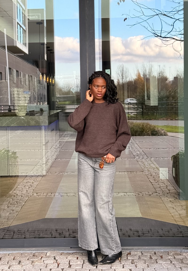 Pull en tricot marron, jeans gris à jambes larges et bottes noires à talons. Debout près d'une entrée en verre avec un décor extérieur en arrière-plan.