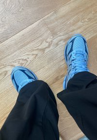 Sapatilhas atléticas em malha azul com detalhes em azul escuro e solas texturizadas, combinadas com calças pretas, posicionadas sobre um chão de madeira.