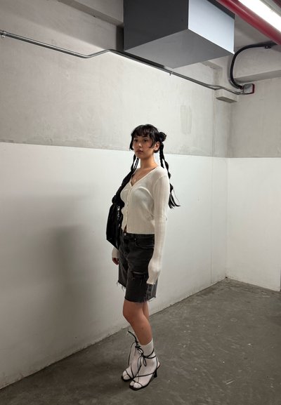 Mujer joven con cabellos trenzados se encuentra en una habitación sencilla vistiendo un cárdigan blanco, una falda negra desgastada, calcetas blancas y tacones destalonados.