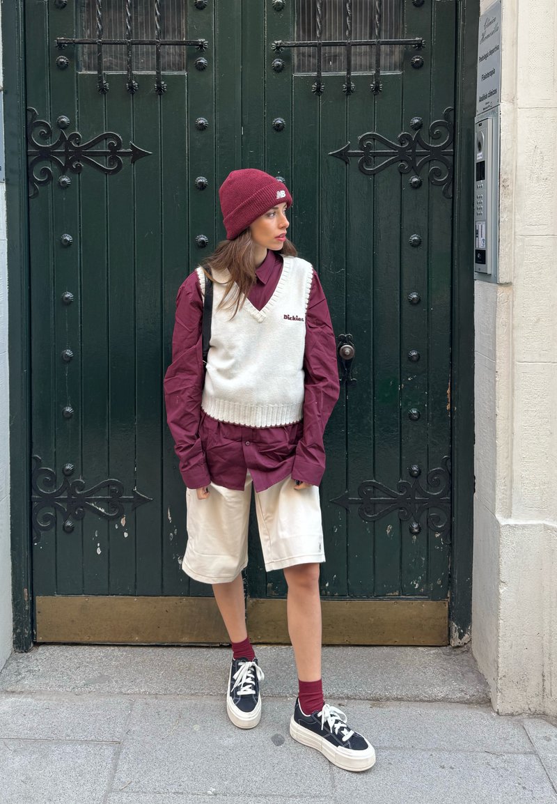 Jeune personne portant une chemise bordeaux, un gilet en tricot blanc, un short beige, un bonnet bordeaux, des chaussettes assorties et des baskets noires, debout devant une porte vert foncé.