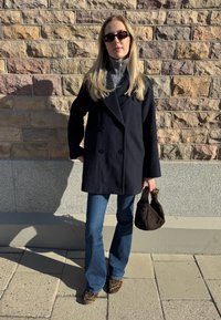 Juodas dvispalvis paltas, pilkas marškiniai su aukštu kaklu, mėlyni platūs džinsai, rudas pintas rankinės ir leopardų raštu batai prieš akmenų sieną.