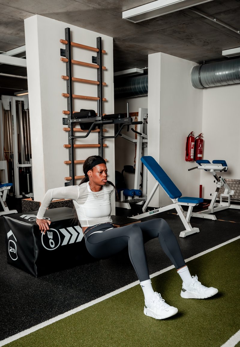 Femme en tenue de sport faisant des dips triceps en utilisant une boîte rembourrée dans une salle de sport avec des machines de musculation et des bancs en arrière-plan.