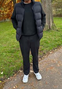 Veste matelassée noire portée sur un pull noir, associée à un pantalon noir et des chaussures de sport gris clair, sur fond vert en plein air.