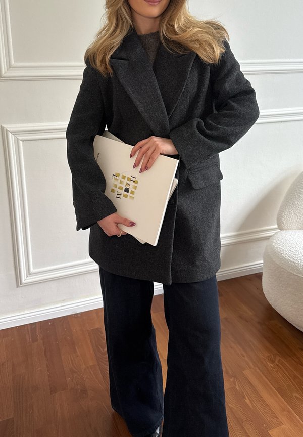 Donna con capelli biondi che indossa un cappotto grigio scuro e pantaloni neri, che tiene in mano una cartella bianca con motivo geometrico oro e nero.