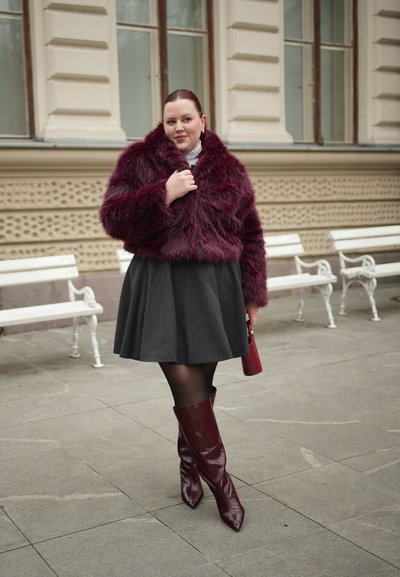 Mujer vestida con una chaqueta de piel sintética burdeos, falda negra y botas burdeos hasta la rodilla, sosteniendo un bolso a juego, de pie sobre una acera de piedra.