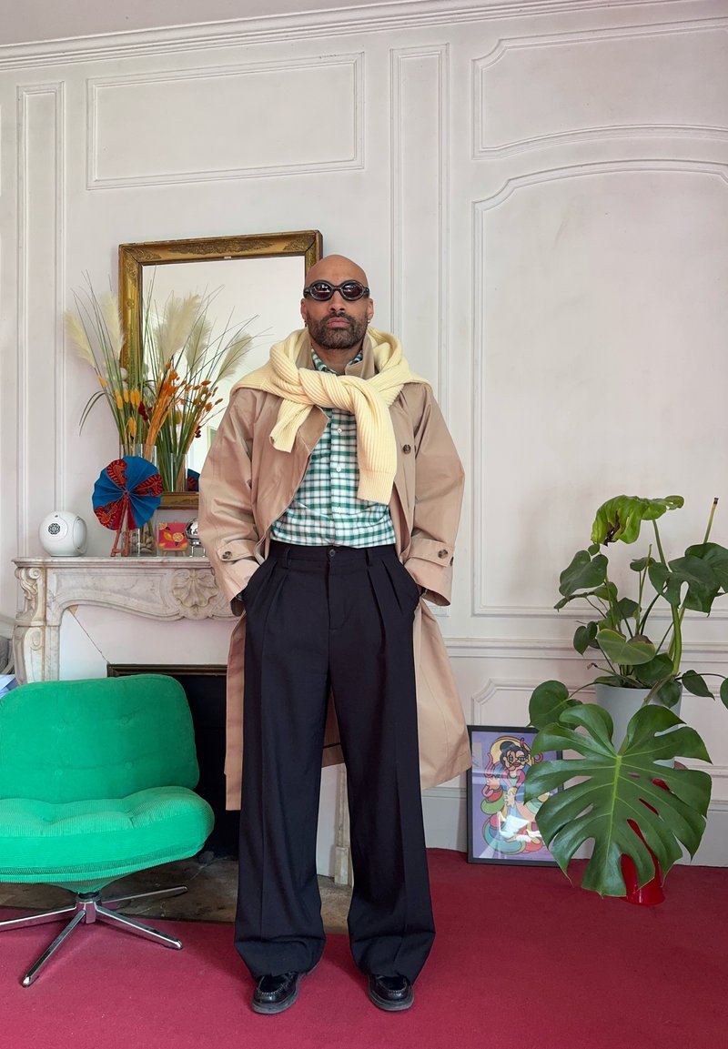 Homme portant des lunettes de soleil, un trench-coat beige, une chemise à carreaux et un pull jaune drapé sur les épaules, debout dans une pièce avec des plantes et des meubles.